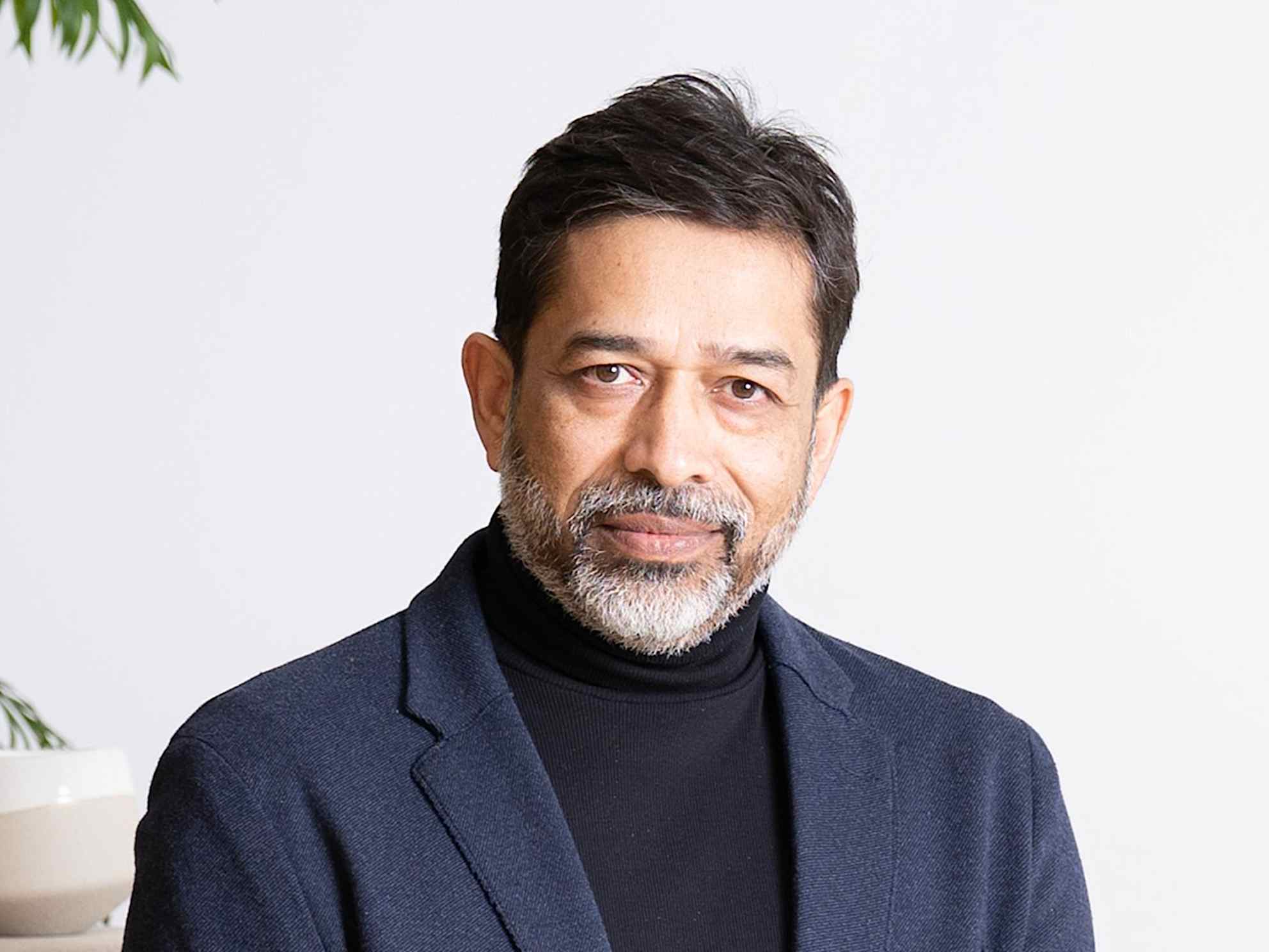Professional headshot of Asheesh Saksena wearing a black turtleneck shirt underneath a navy-blue sport coat. 